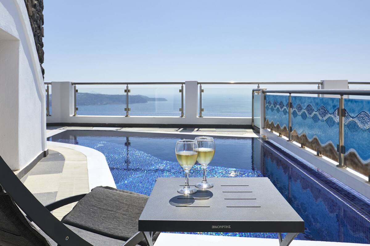 A poolside area with two glasses of white wine on a table, overlooking a clear blue sea.
