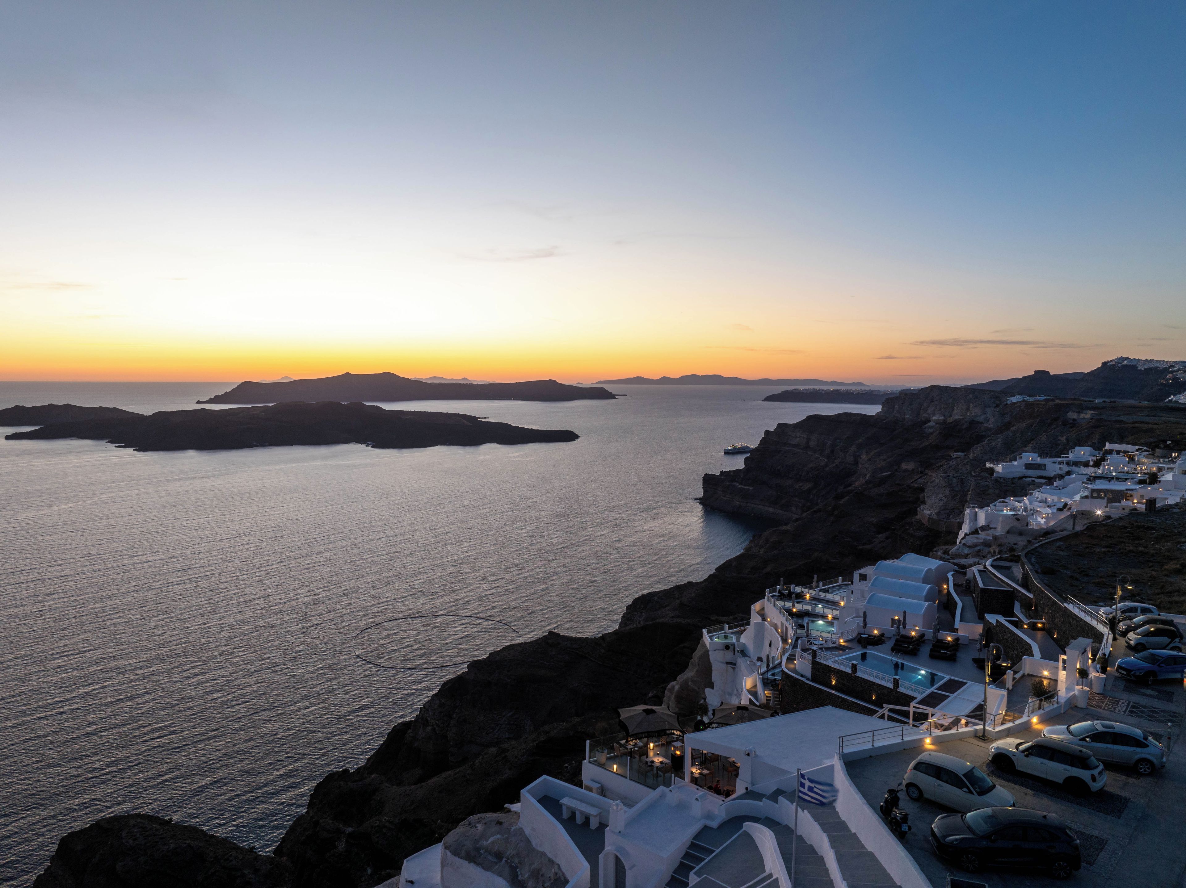 A coastal hotel with white buildings, a marina, and a view of the sea and islands at sunset.
