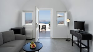 A modern living room with a grey sofa, a small round table with fruit, a TV on a black stand, and a view of the sea through open doors. - Petit Palace (Santorini)