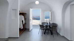 A room with a dining table and chairs, a view of the sea through an open door, and a white robe hanging on a rack. - Petit Palace (Santorini)
