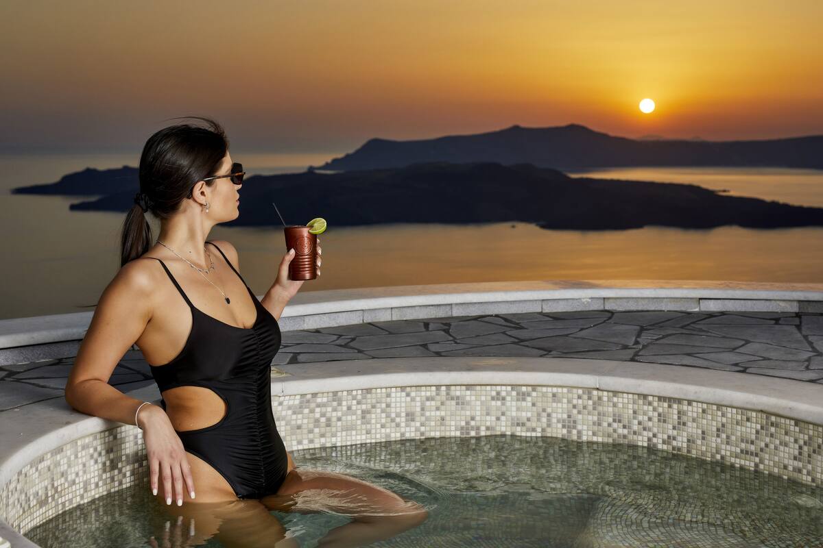 A person in a black swimsuit sitting by a pool with a sunset view.