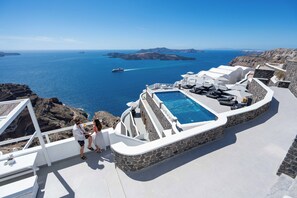 A pool area with a view of the sea and islands, surrounded by white architecture and stone walls. - Petit Palace (Santorini)