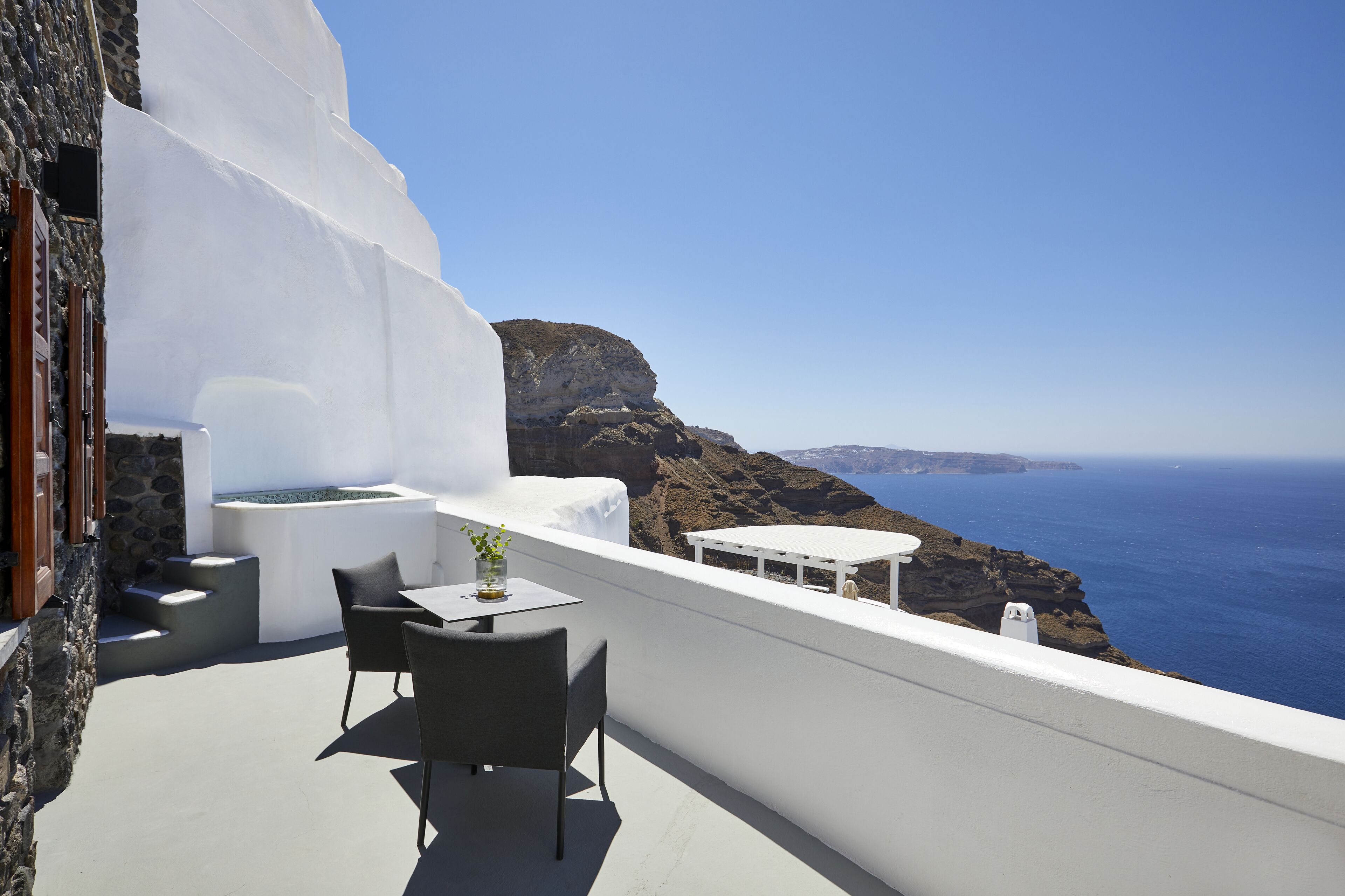 A white-walled balcony with a table and chairs, overlooking a coastal landscape with cliffs and the sea.