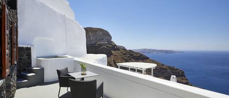 A white-walled balcony with a table and chairs, overlooking a coastal landscape with cliffs and the sea.