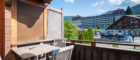 A wooden balcony with a table and chairs, overlooking a town with a large hotel and mountains in the background.