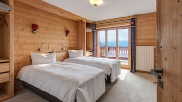 A wooden cabin bedroom with two beds, a wooden wardrobe, and a window with a view of mountains.