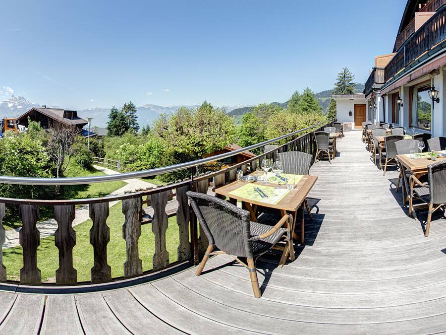 Una terraza de madera con asientos exteriores, vista a montañas y un edificio con balcón.
