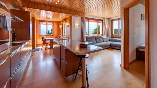 A modern kitchen with wooden cabinetry and a central island, a cozy seating area with a sofa and a dining table, and large windows offering a view of the outdoors. - Alpe Fleurie (Ollon)