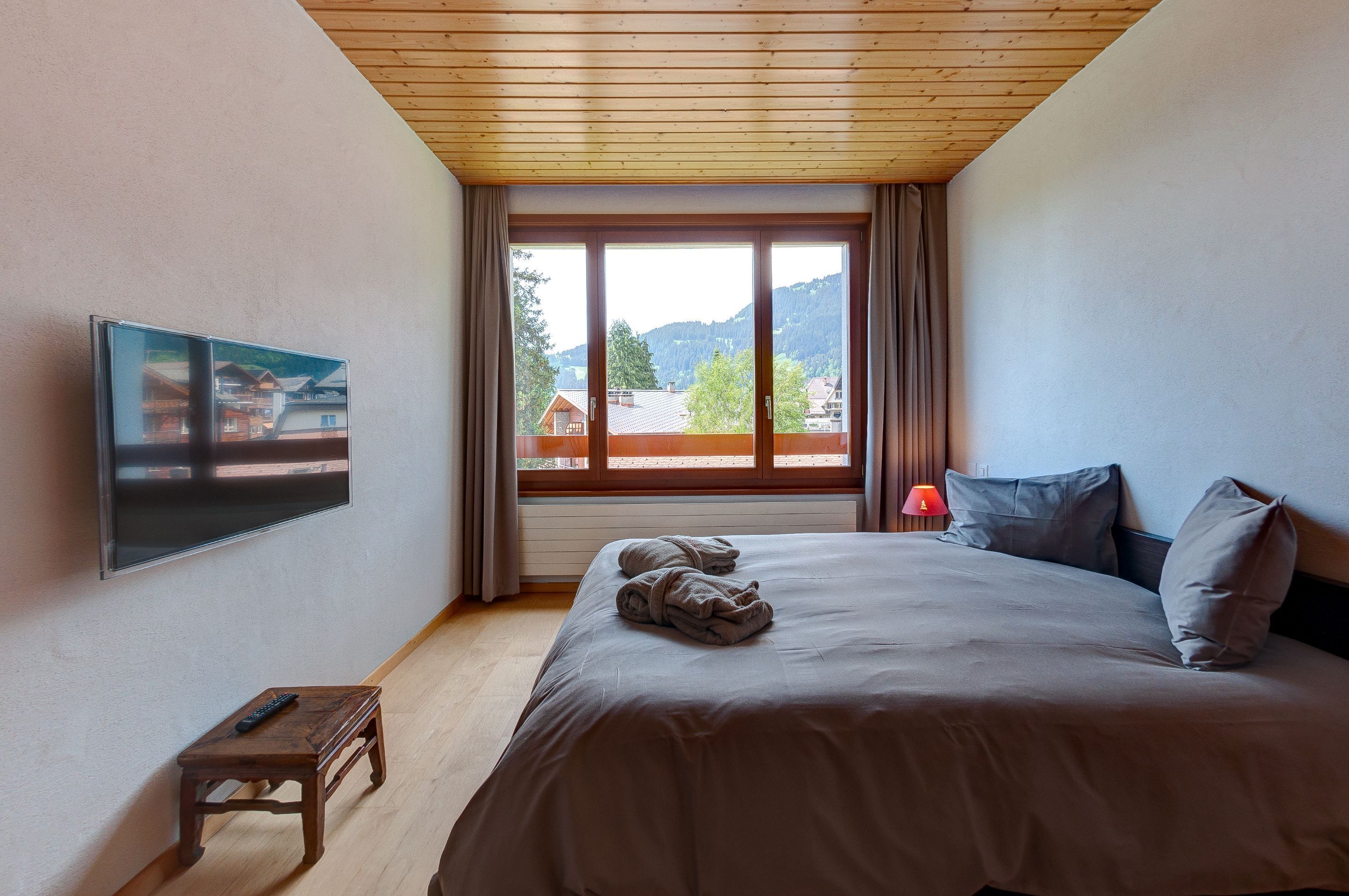 Un dormitorio con una cama grande, un televisor montado en la pared, una mesita con un control remoto y una ventana con cortinas.