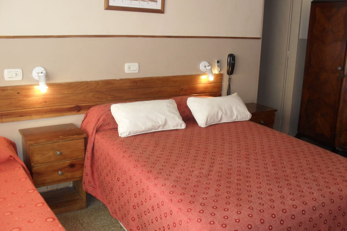 A hotel room with a wooden headboard, two bedside tables with lamps, a bed with a red patterned bedspread, and a framed picture on the wall.