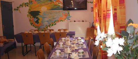 Un comedor con una mesa larga dispuesta para una comida, sillas alrededor, un televisor en la pared y un cuadro decorativo sobre la mesa.