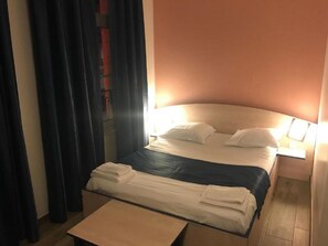 Una habitación de hotel con cama, mesita de noche y ventana con cortinas.
