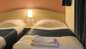 A hotel room with two beds, each with white linens and a folded towel on the bed. There is a nightstand with a lamp between the beds.