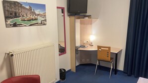 A hotel room with a red chair, a desk with a lamp, a TV, and a picture of a fountain on the wall.