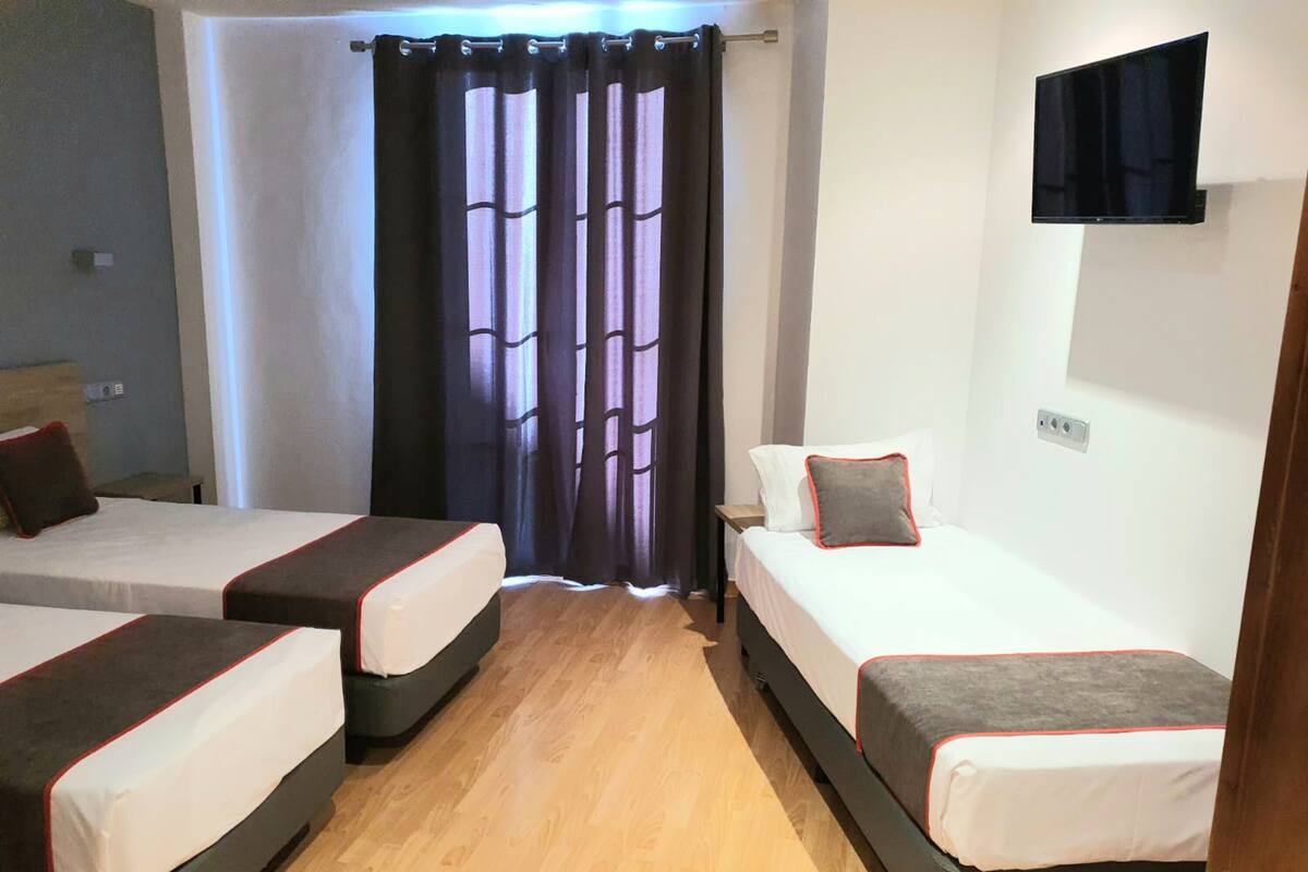 Hotellihuone, jossa on kolme erillistä sänkyä, seinälle asennettu taulutelevisio ja ikkuna, jossa on verho.