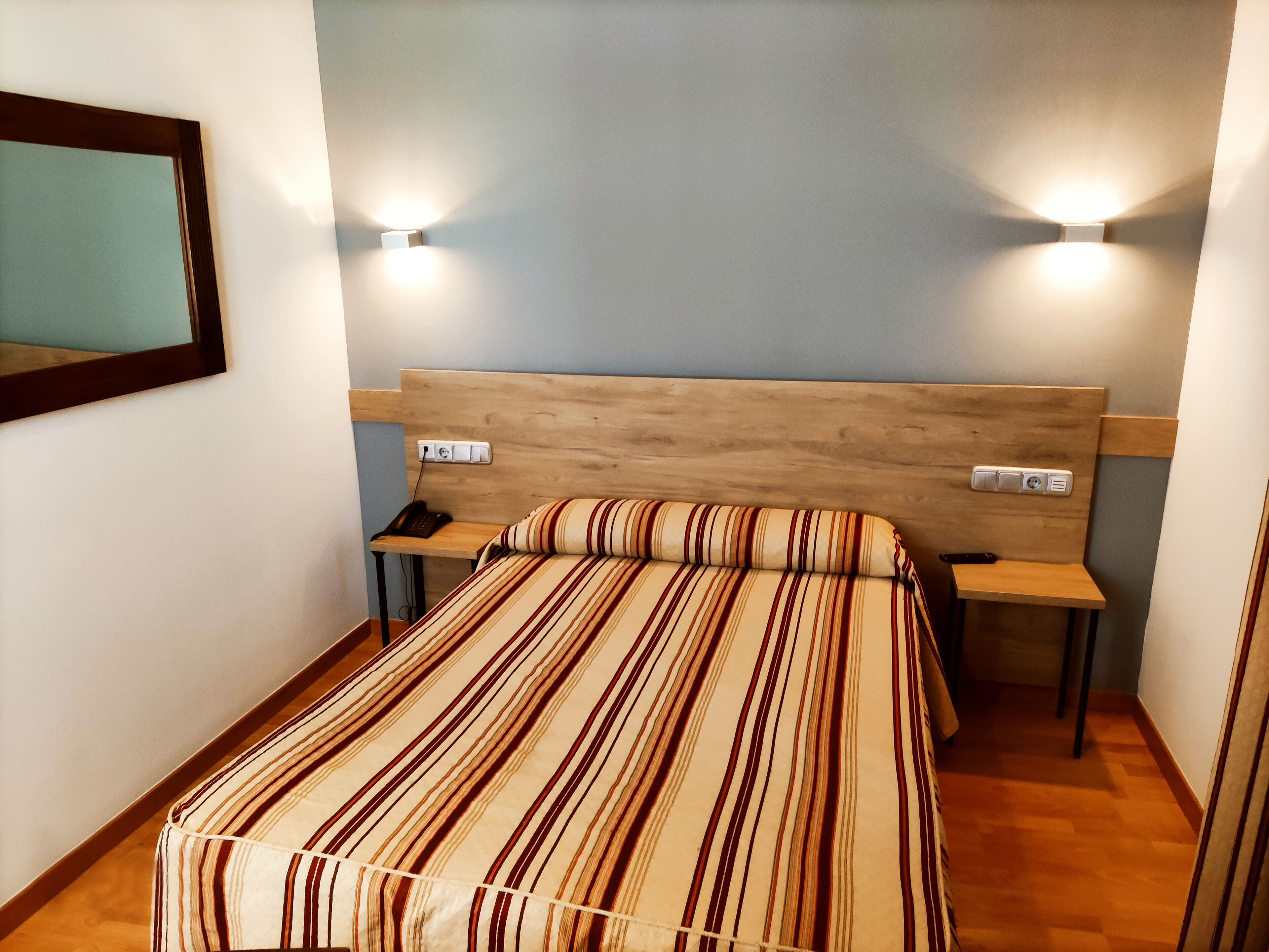 A bedroom with a striped bedspread, two bedside tables with lamps, a wooden headboard, and a wall-mounted mirror.