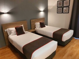 Dos camas individuales con cabeceras de madera, ropa de cama blanca con detalles rojos y una lámpara fijada a la pared.