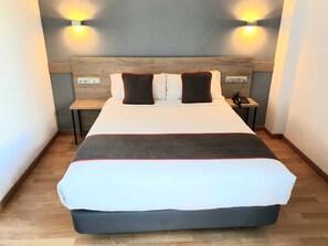 Una cama bien hecha con edredón gris y blanco, dos mesitas de noche con lámparas y un cabecero de madera.