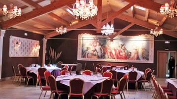 A banquet hall with high wooden ceilings, chandeliers, and red upholstered chairs around tables set for dining.