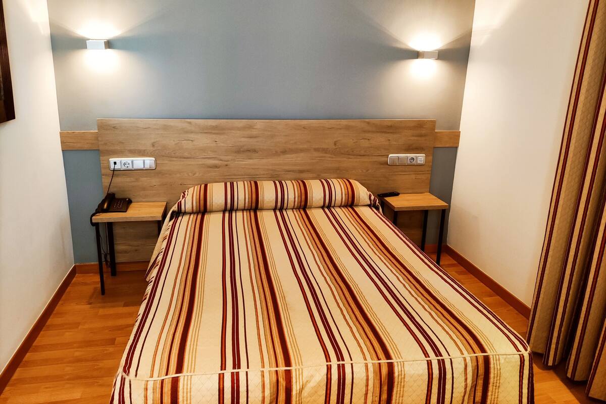 Hotellihuone, jossa on puinen sängynpääty, raidallinen päiväpeite, yöpöydät lampuineen ja pistorasia.