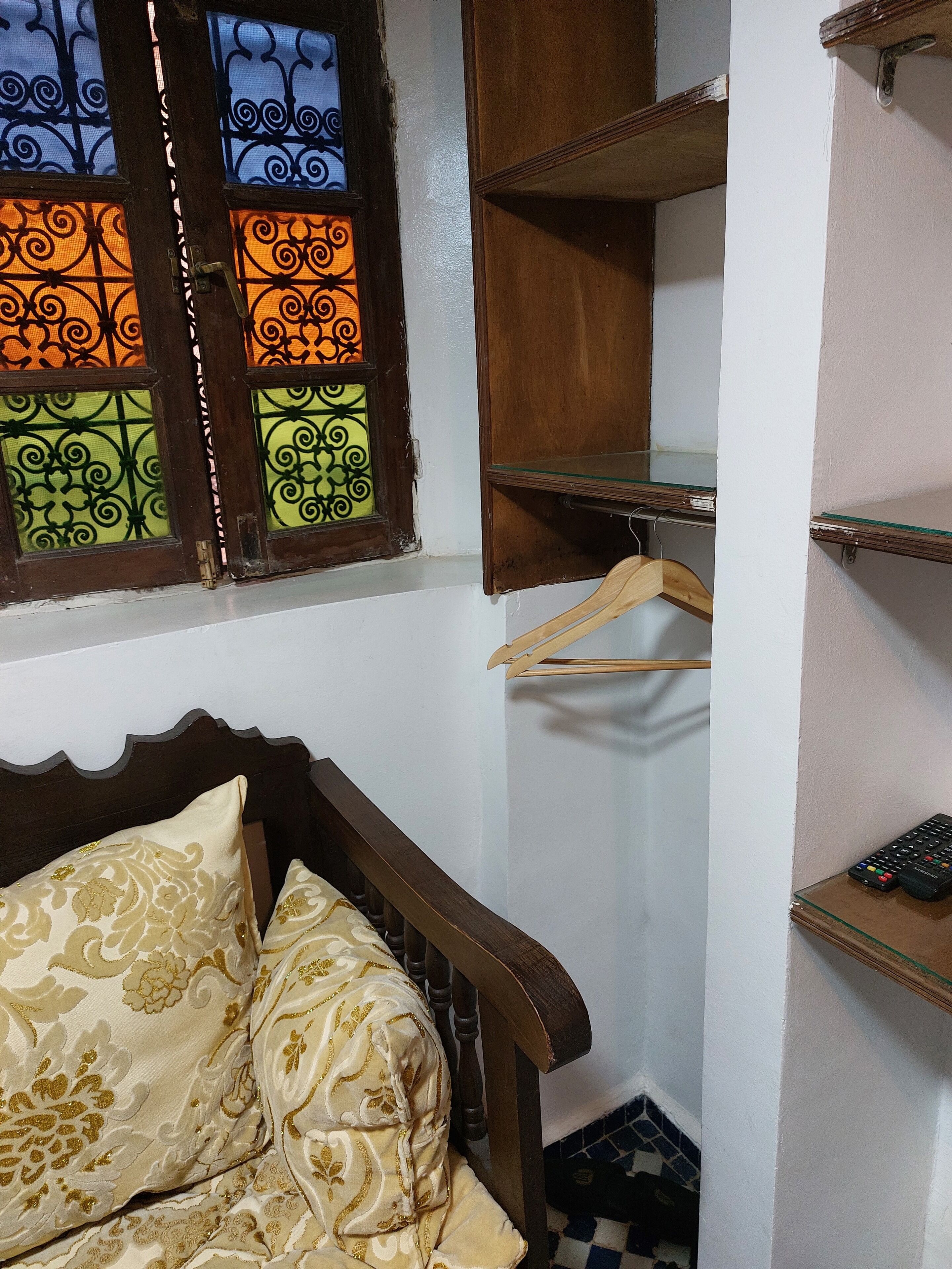 A bedroom with a wooden bed, a window with decorative glass panes, and a small shelf with a remote control.