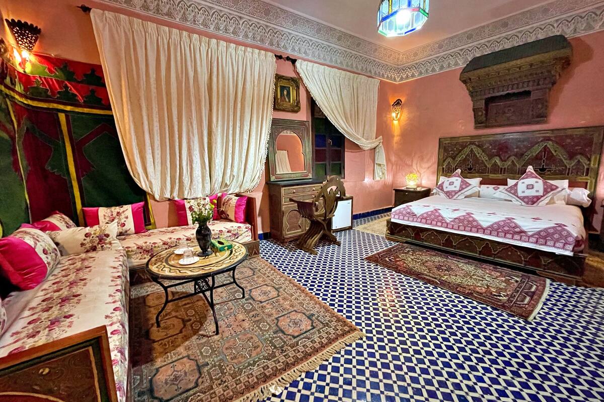A traditional Moroccan room with a checkered floor, a sofa with floral cushions, a bed with patterned bedding, and a decorative mirror.