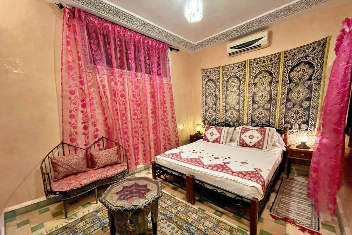 A room with a bed, a chair, a small table, patterned curtains, and a decorative wall tapestry.