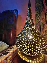Luminária decorativa com padrões recortados detalhados, colocada sobre uma superfície de madeira e com um fundo de tecido estampado.