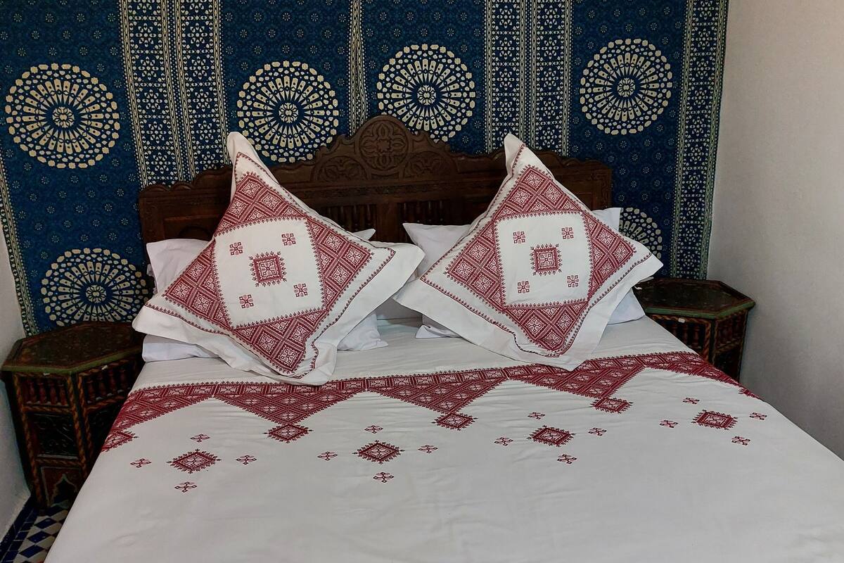 A bed with white and red patterned bedding, a wooden headboard, and a blue patterned tapestry on the wall.