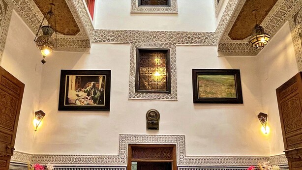 Un comedor con techos altos, patrones ornamentales en el techo y un acceso central. Hay dos mesas redondas dispuestas con manteles blancos, cojines azules y cubiertos. Las paredes presentan intrincados trabajos en azulejos y obras de arte enmarcadas.