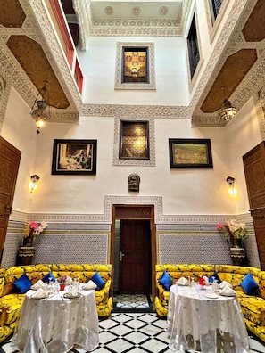A dining area with a high ceiling, ornate ceiling patterns, and a central doorway. There are two round tables set with white tablecloths, blue cushions, and silverware. The walls feature intricate tile work and framed artwork. - Riad Dar Guennoun (Fes)