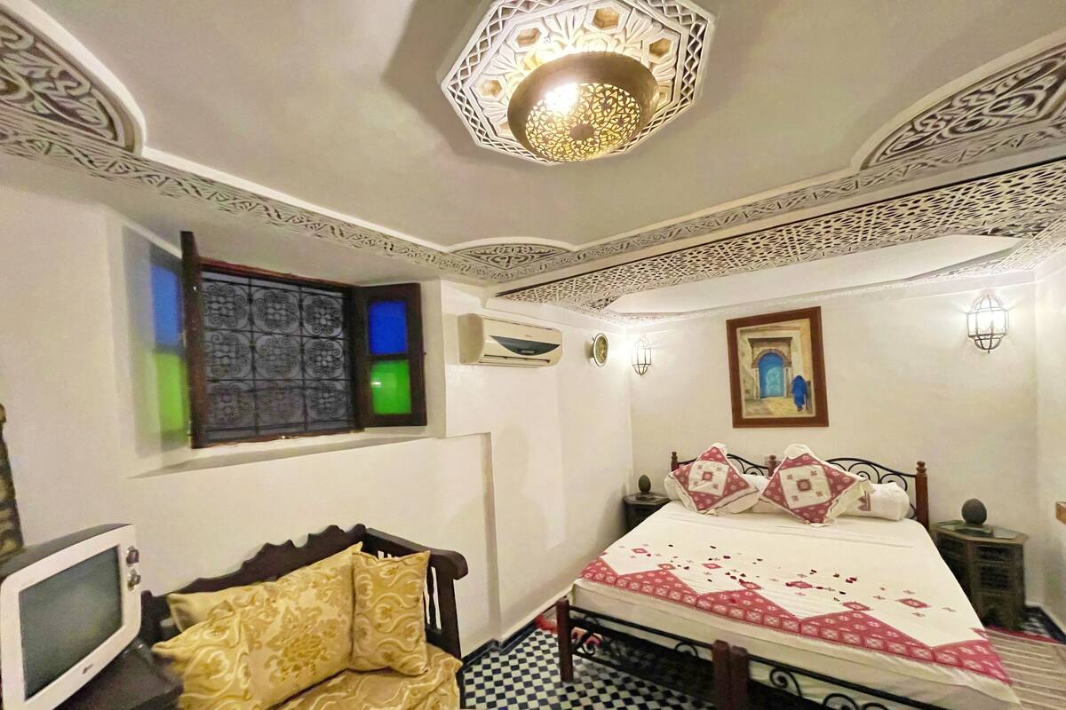 A bedroom with a bed, a chair, a television, a window with patterned glass, and decorative ceiling and wall elements.