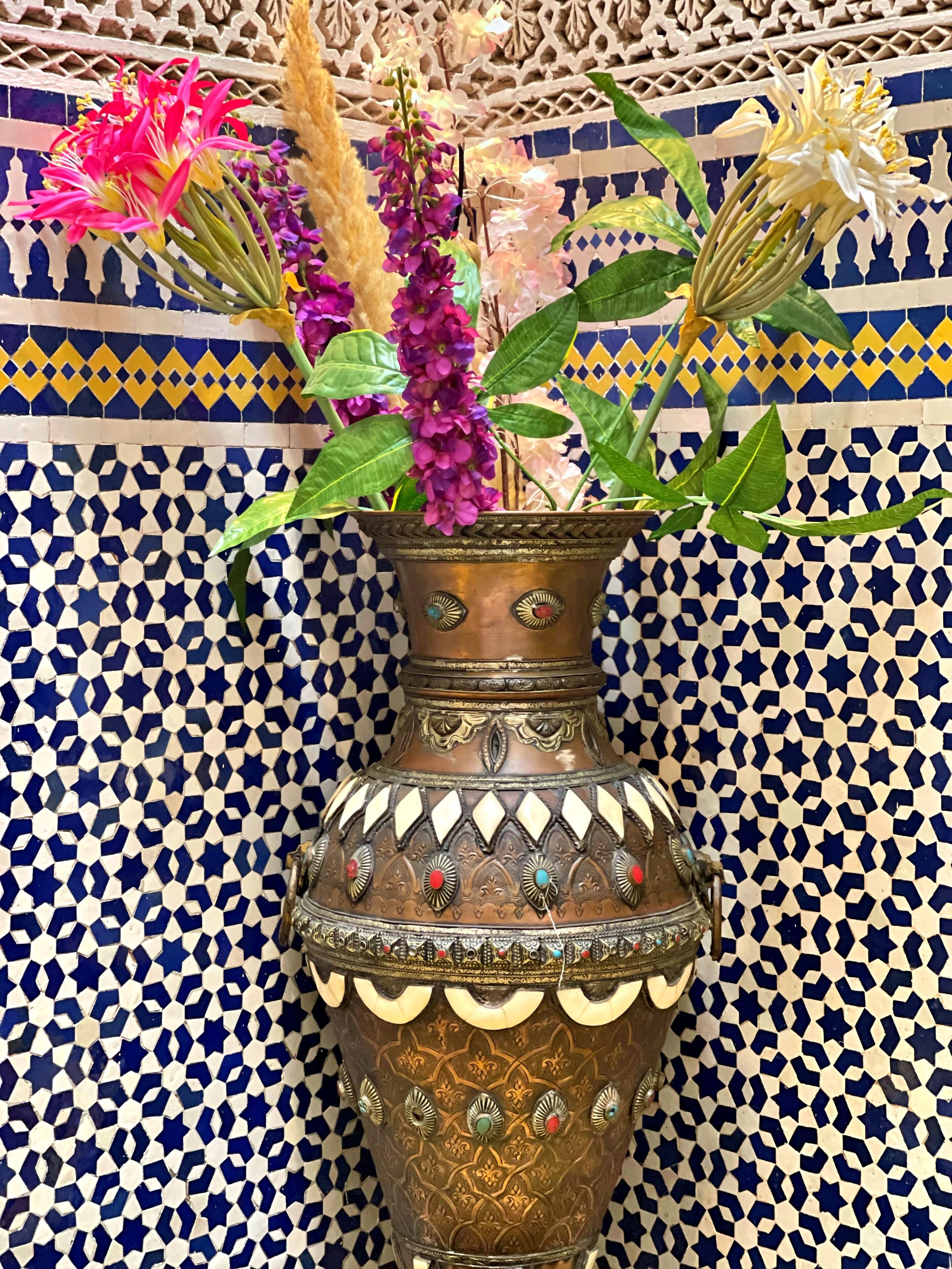 A decorative vase with intricate patterns and colorful flowers against a tiled wall with geometric designs.