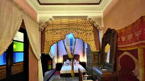 Una habitación tradicional con cama, mesita de noche y silla. Las paredes presentan un diseño decorativo y cuenta con un arco ornamental.