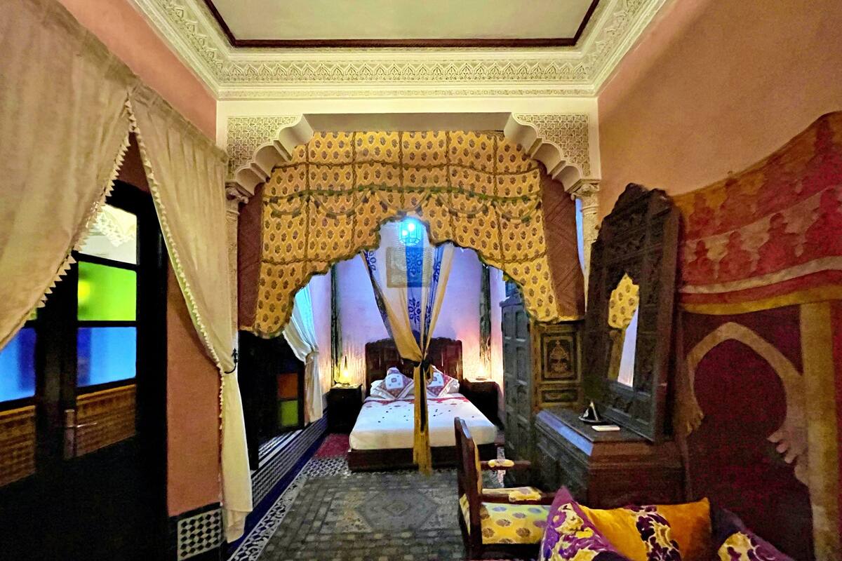 A traditional room with a bed, a nightstand, and a chair. The room has patterned walls and a decorative archway.