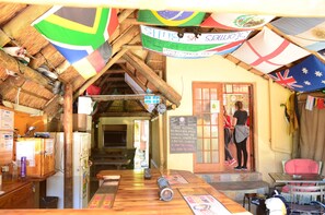 A rustic bar with wooden furniture and a thatched roof. Flags are hung from the ceiling, and a chalkboard sign is visible. - A Sunflower Stop - Hostel (Cape Town)