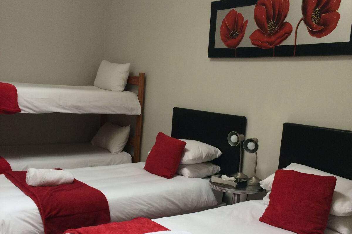 Ein Hotelzimmer mit drei Betten, jeweils mit weißer Bettwäsche und roten Akzenten. Über den Betten hängt ein gerahmtes Kunstwerk und dazwischen steht ein kleiner Tisch mit einer Lampe.
