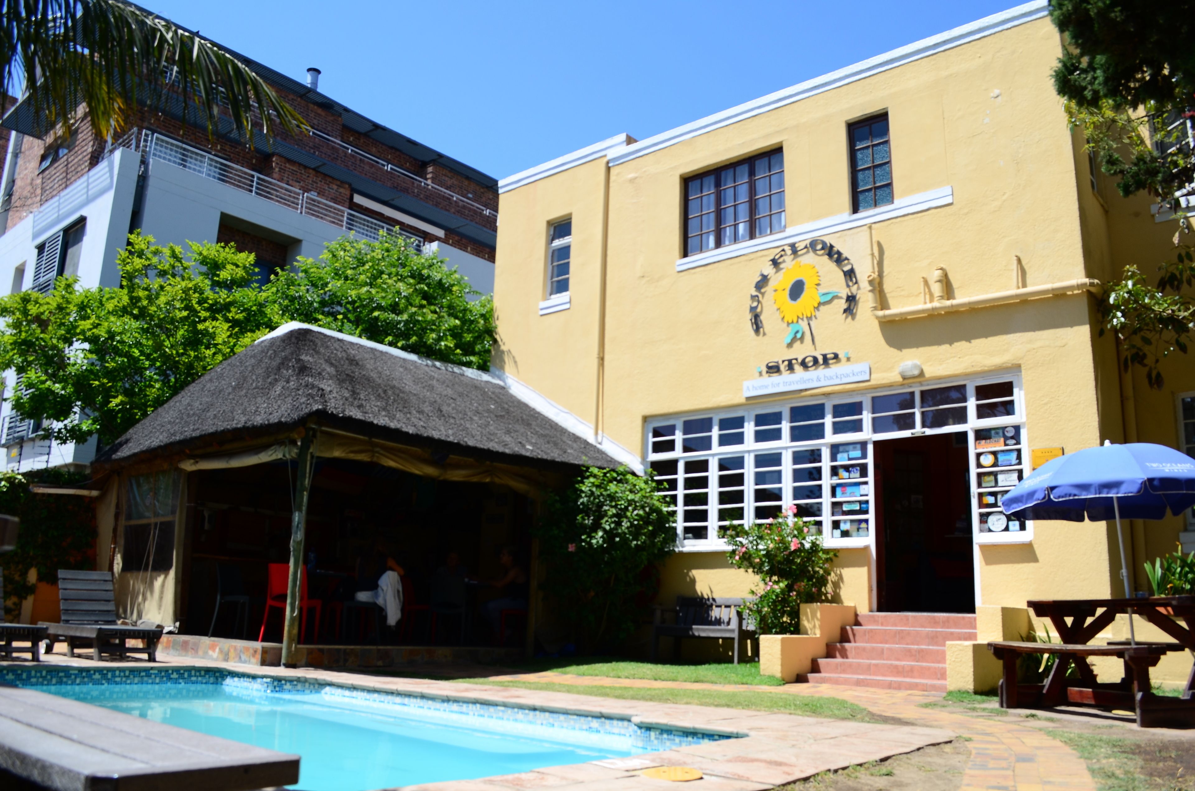 A building with a sun emblem and the word "STOP" on the facade, a pool area with a thatched-roof structure, and outdoor seating.