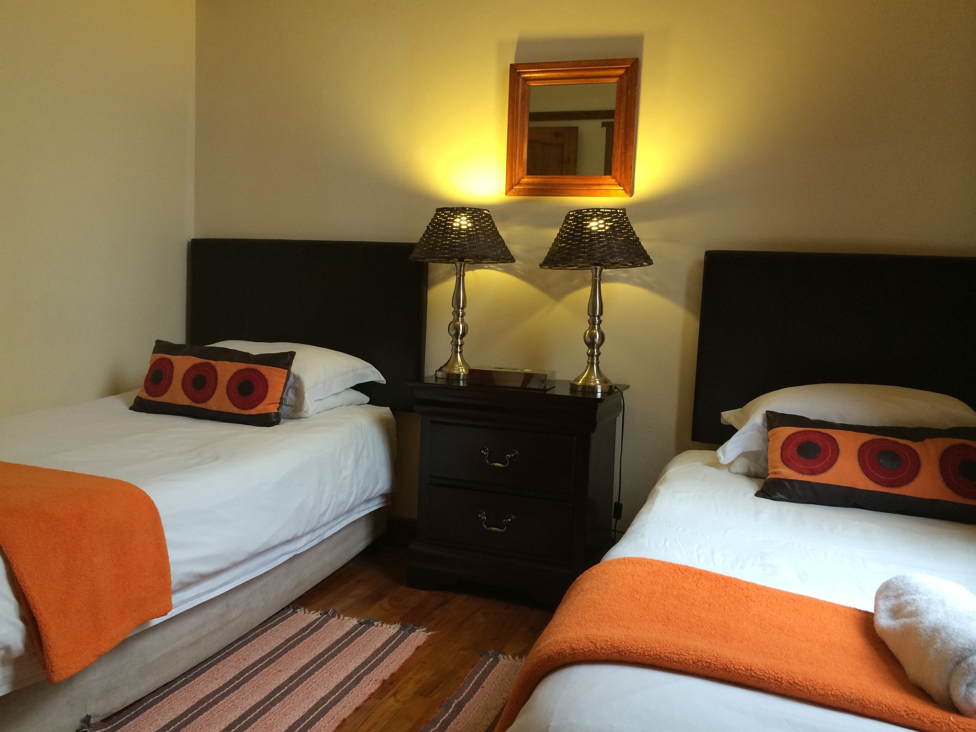 A hotel room with two single beds, each with a white duvet and orange pillows. There are two bedside tables with lamps, a wooden headboard, and a mirror on the wall.