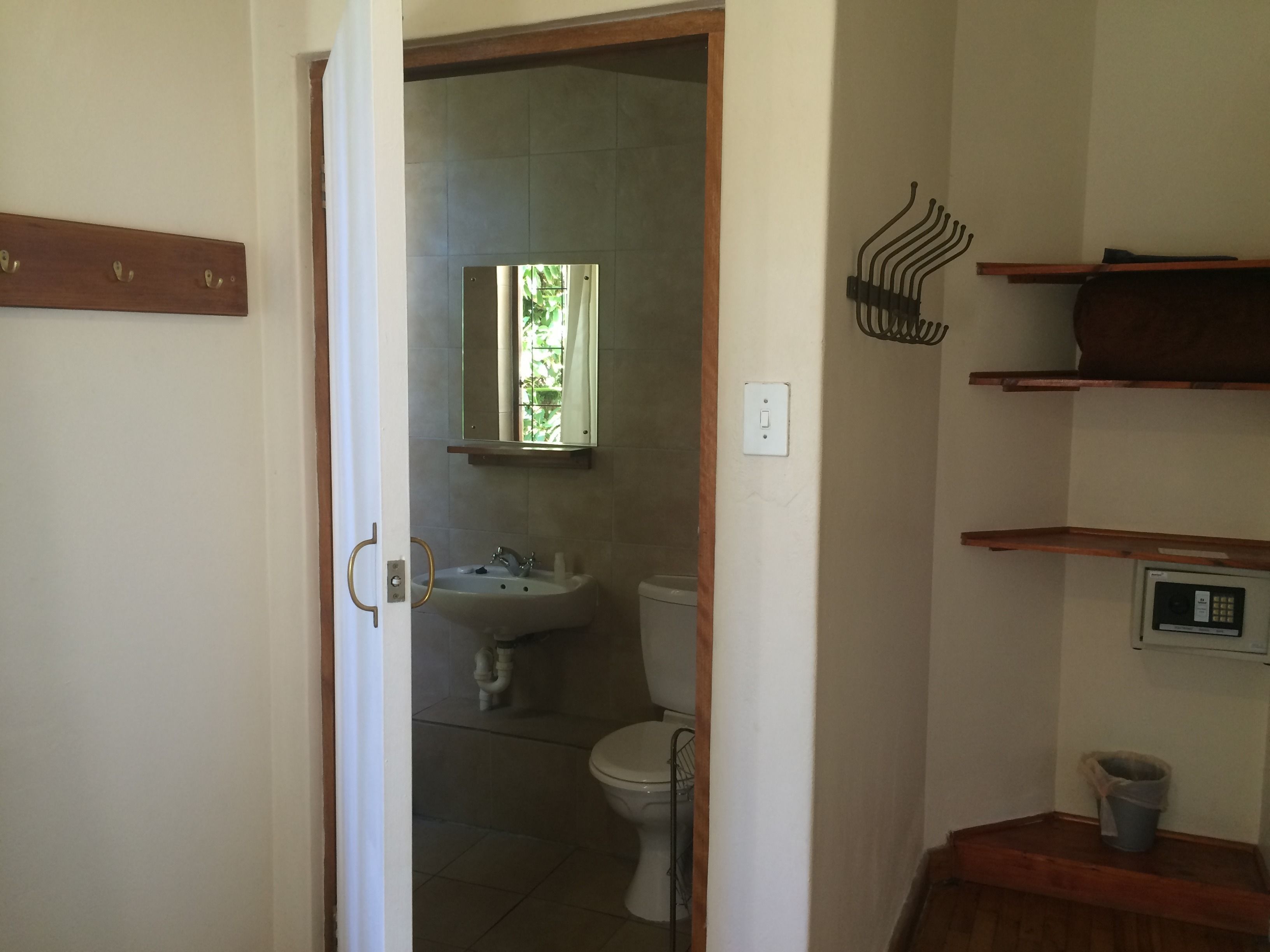 Une salle de bain avec des toilettes, un lavabo et un miroir. Il y a des étagères en bois au-dessus du lavabo et une poubelle à droite.