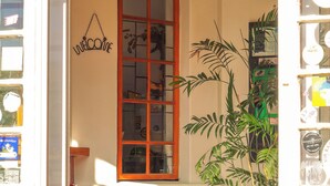 A doorway leading to a room with a red cushion on a black sofa, a wooden door with a glass panel, a potted plant, and a welcome mat. - A Sunflower Stop - Hostel (Cape Town)