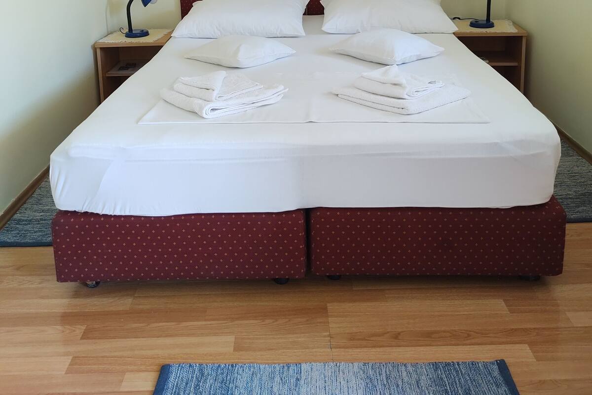 A double bed with white linens and red bedding, two bedside tables with lamps, a framed painting on the wall, and an air conditioning unit.