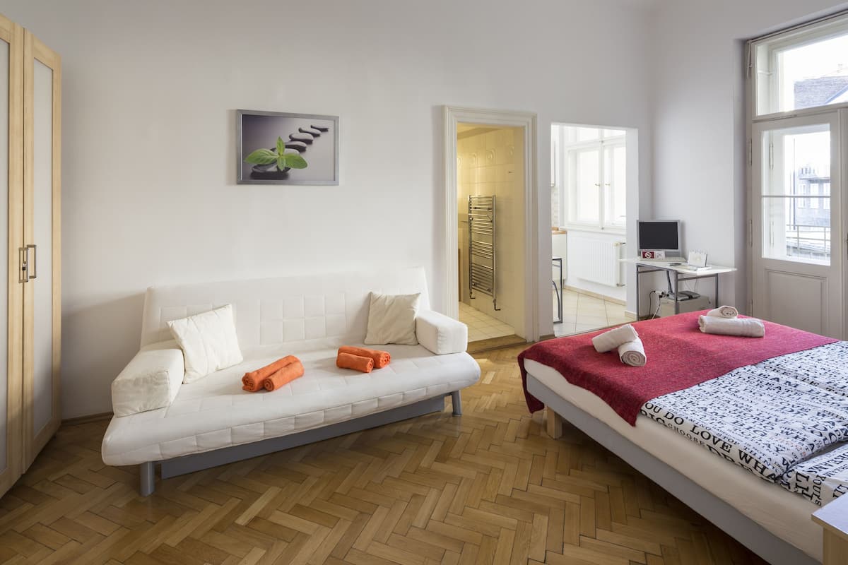 Egy modern szállodai szoba, amelyben van egy kanapé, egy piros takaróval borított ágy, egy íróasztal számítógéppel, és a városra néző kilátás nyílik az ablakon keresztül.