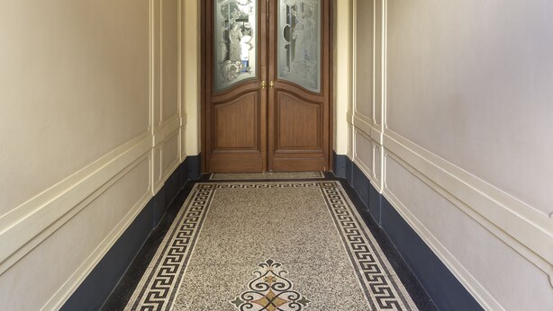 Um corredor com detalhes ornamentais no teto, uma porta dupla de madeira com painéis de vidro e piso estampado.