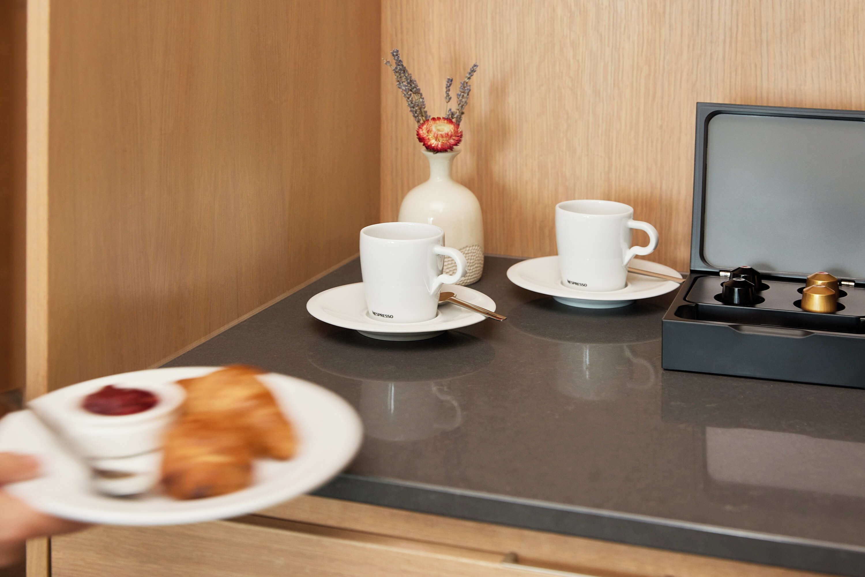 A breakfast setup with two white cups, a plate with croissant and jam, and a vase with dried flowers.