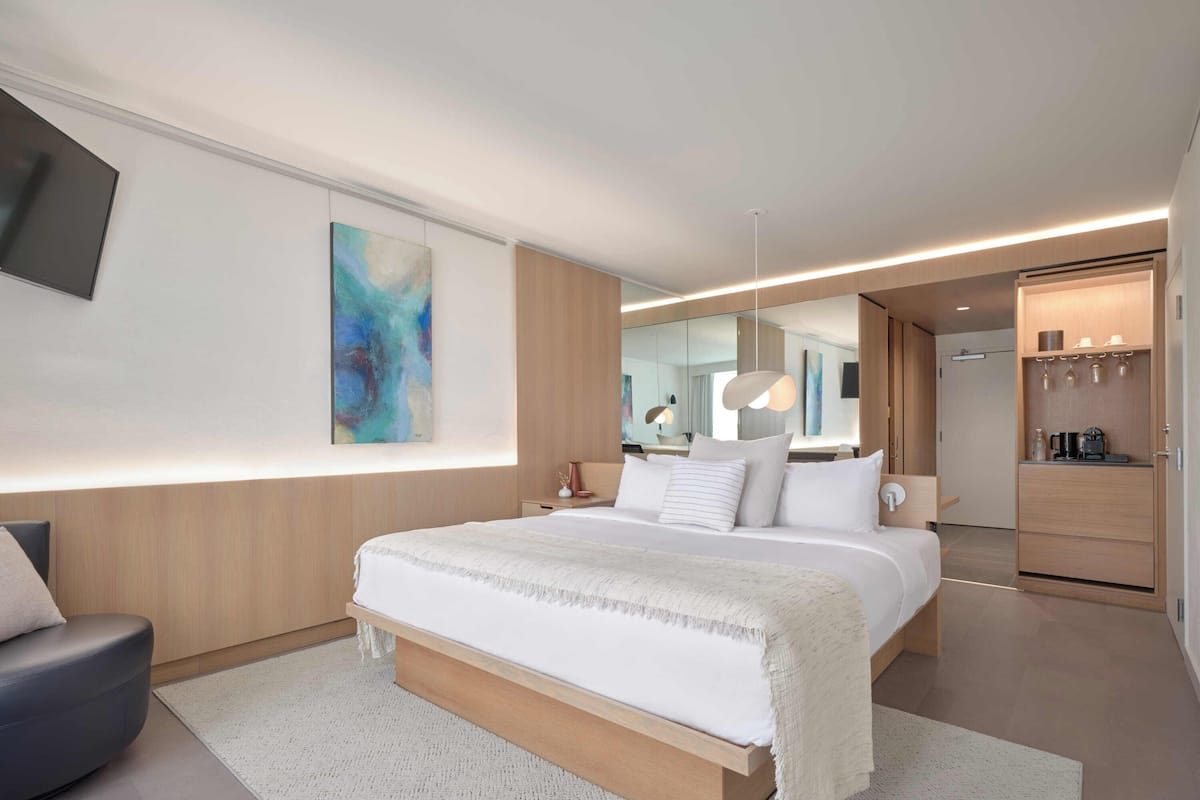 A modern hotel room with a large bed, a seating area, a coffee machine, and a large window.