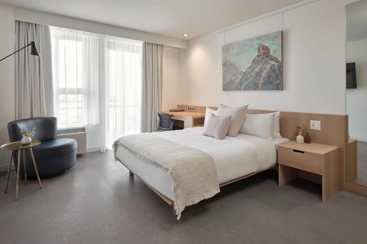 A modern hotel room with a large bed, a desk, a chair, and a painting on the wall.