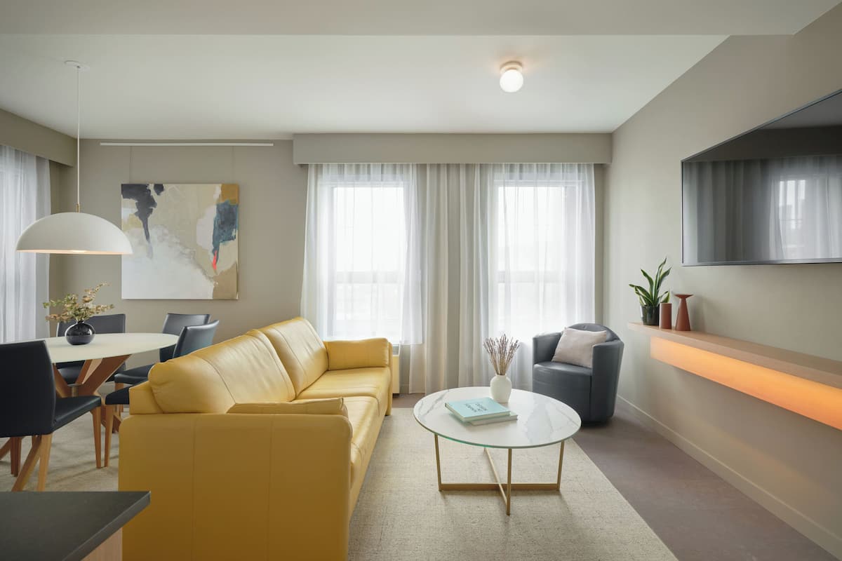 A modern living room with a yellow sofa, a glass coffee table, and a dining area with a white table and chairs.