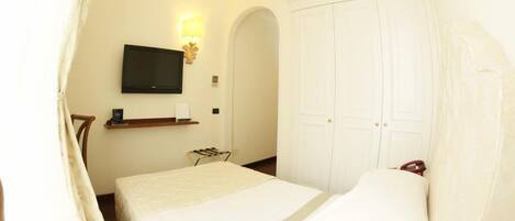Habitación de hotel con cama, televisor montado en la pared, armario y una pequeña mesa con lámpara.