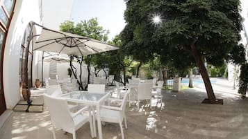 Un patio al aire libre con mobiliario de mimbre blanco y un gran paraguas, rodeado de árboles y una piscina.